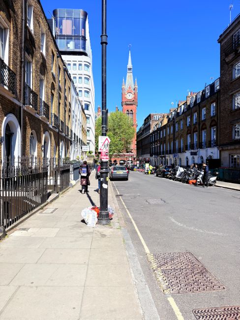 A city street in Fulham during daytime with rows of brick residential buildings on both sides, featuring large windows and front gardens. The sidewalk is clean with a few pedestrians, some carrying shopping bags. A black lamppost stands near the curb, and parked vehicles, including motorcycles and small cars, line the street. In the background, the prominent clock tower of a historic church with a tall spire and red brick façade is visible against a bright blue sky. The scene is well-lit with natural sunlight, highlighting the tidy and well-maintained surfaces of the buildings and street, reflecting a typical urban residential area in Fulham.