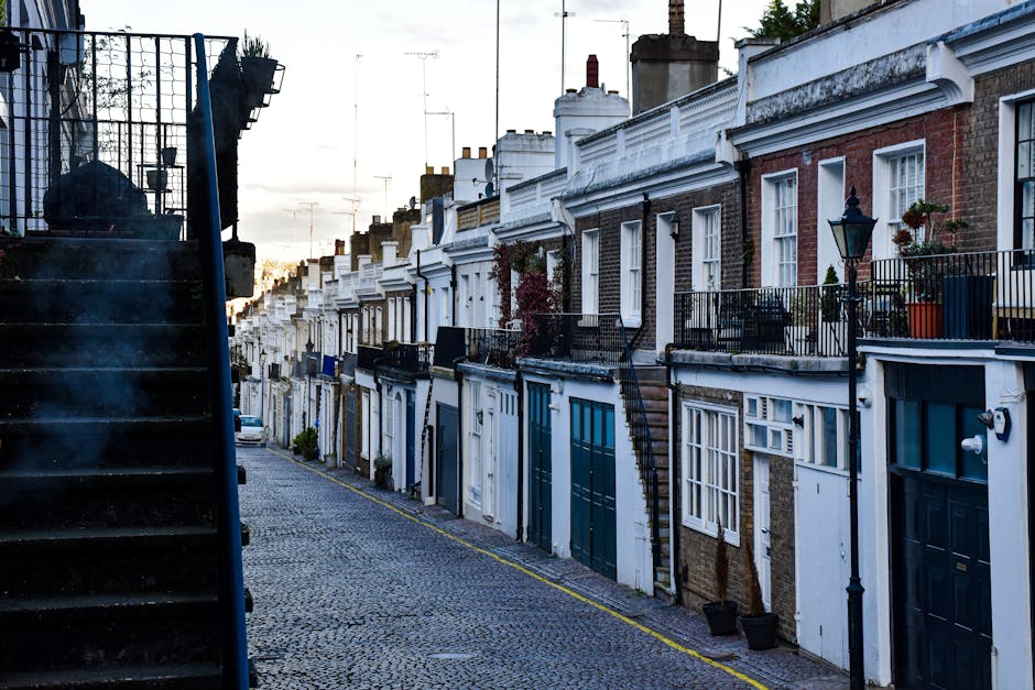 A row of traditional terraced house facades along a cobbled street in Fulham, London, featuring brick exterior walls and white-painted decorative moldings. Small balconies with black iron railings are visible on some upper levels, with potted plants placed on them. The street is illuminated by vintage-style street lamps, and the scene is captured during a clear day with natural light emphasizing the cleanliness and well-maintained condition of the exteriors. The image showcases the external surfaces of residential buildings, highlighting brickwork and painted wood elements, consistent with typical residential architecture in Fulham. Fulham Carpet Cleaning may utilize surface cleaning and deep cleaning techniques to maintain the appearance and hygiene of similar residential areas.