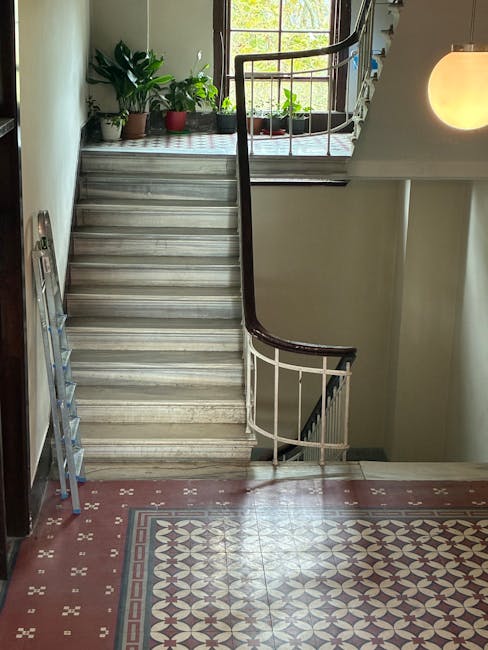 Interior view of a residential staircase in a narrow flat, showing a marble or stone staircase with a dark wooden handrail and white metal balustrade. The staircase leads up to a sunlit landing with a