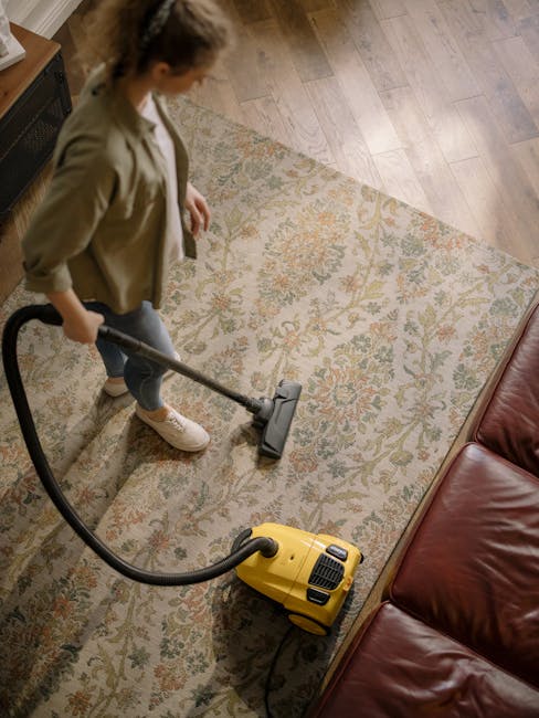 A person using a yellow canister vacuum cleaner to deep clean a patterned area rug in a living room. The rug features a floral design with beige, green, and orange tones, placed on a wooden floor. The individual is wearing a beige jacket and jeans, focused on surface cleaning, with natural light illuminating the room. Nearby, a brown leather sofa is partially visible, and the scene emphasizes thorough upholstery and carpet hygiene, representing domestic cleaning services provided by Fulham Carpet Cleaning in Fulham.