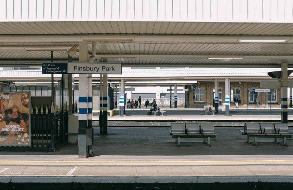 The image depicts Fulham Broadway train station platform with a covered seating area. The platform surface is made of concrete, appearing clean and well-maintained. Several metal benches are arranged parallel to the platform edge, with a few passengers seated and waiting for the train. Overhead, the station roof is supported by metal pillars painted with blue and white stripes. In the background, additional tracks and a station building with large windows are visible. A sign indicates the station's name, 'Finsbury Park,' suggesting nearby transport connections. The lighting is natural, with daylight illuminating the area, highlighting the tidy and hygienic appearance of the station. This scene aligns with professional cleaning standards, emphasizing surface cleanliness and order, relevant to services like Fulham Carpet Cleaning for ensuring hygienic environments.