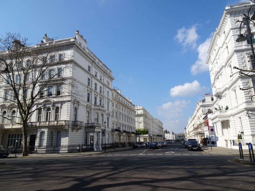 A wide-angle view of a street in Fulham with elegant white residential buildings featuring ornate architectural details and large windows. The buildings are multi-story with balconies, some with metal railings and decorative moldings. The street is paved with asphalt, lined with parked cars, and bordered by leafless trees casting shadows on the road. Bright daylight illuminates the scene with a clear blue sky and scattered white clouds, highlighting the cleanliness and well-maintained facade of the buildings. The scene reflects an urban environment in Fulham, captured in a natural and realistic style, with no visible signage or people. This image exemplifies a well-maintained residential area suitable for surface and deep cleaning services, such as those offered by Fulham Carpet Cleaning.