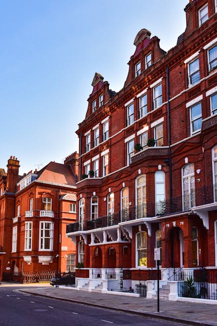 A row of red-brick residential buildings on a street view, featuring large sash windows with white frames, decorative stonework, and black wrought-iron balconies. The ground floor has brick steps leading to entrances, with some steps and doorways visible. The pavement is clean, with a streetlamp and a few parked cars nearby. Bright natural daylight illuminates the scene, highlighting the building's exterior and the clear blue sky above. This image relates to surface and deep cleaning of residential exteriors, as part of domestic cleaning services offered by Fulham Carpet Cleaning. The buildings' surfaces appear well-maintained and free of dust or grime, emphasizing hygiene and maintenance in Fulham flats, consistent with the Lillie Road end of tenancy cleaning service described on the page.