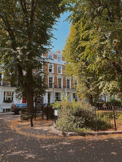 A wide-angle view of a street in Fulham with elegant white residential buildings featuring ornate architectural details and large windows. The buildings are multi-story with balconies, some with metal railings and decorative moldings. The street is paved with asphalt, lined with parked cars, and bordered by leafless trees casting shadows on the road. Bright daylight illuminates the scene with a clear blue sky and scattered white clouds, highlighting the cleanliness and well-maintained facade of the buildings. The scene reflects an urban environment in Fulham, captured in a natural and realistic style, with no visible signage or people. This image exemplifies a well-maintained residential area suitable for surface and deep cleaning services, such as those offered by Fulham Carpet Cleaning.