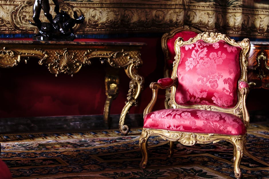 A luxurious Victorian-style armchair upholstered in vibrant pink damask fabric with intricate floral patterns, featuring a carved wooden frame with gilded detailing and curved legs. The chair is positioned on a richly patterned carpet with floral motifs, in front of a marble-topped side table that has ornate, gold-accented carvings. The background reveals a deep red wall and an elaborate, decorative painting or tapestry with gold and bronze tones. The setting appears well-maintained with no visible dust or stains, exemplifying high-end interior decor suitable for deep surface cleaning or domestic maintenance by Fulham Carpet Cleaning as part of their upholstery cleaning services for period homes in Fulham.