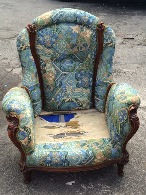 A vintage-style upholstered armchair with wooden carved arms and legs, situated outdoors on a paved surface. The fabric features a faded blue, green, and beige floral pattern, with visible tears and worn areas, especially on the seat cushion where the stuffing and underneath support are exposed. The chair appears dusty and aged, highlighting the need for thorough deep cleaning and restoration, which Fulham Carpet Cleaning offers as part of their domestic cleaning services. Natural lighting accentuates the texture of the worn fabric and the intricate woodwork, with the background showing a blurred street scene.