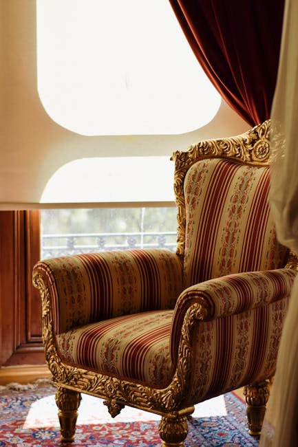 A luxurious Victorian-style armchair upholstered in vibrant pink damask fabric with intricate floral patterns, featuring a carved wooden frame with gilded detailing and curved legs. The chair is positioned on a richly patterned carpet with floral motifs, in front of a marble-topped side table that has ornate, gold-accented carvings. The background reveals a deep red wall and an elaborate, decorative painting or tapestry with gold and bronze tones. The setting appears well-maintained with no visible dust or stains, exemplifying high-end interior decor suitable for deep surface cleaning or domestic maintenance by Fulham Carpet Cleaning as part of their upholstery cleaning services for period homes in Fulham.