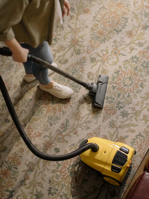A person using a vacuum cleaner with a yellow canister and a black hose to deep clean a floral-patterned carpet in a residential setting. The individual is wearing beige shoes, jeans, and a beige jacket, and is standing on a large, ornate rug with a cream background and muted green, orange, and beige floral motifs. The room is well-lit, and the carpet appears to be in the process of being thoroughly vacuumed by Fulham Carpet Cleaning as part of their domestic cleaning and surface sanitation services. The focus is on the carpet and the cleaning tool, with furniture not visible in the frame.