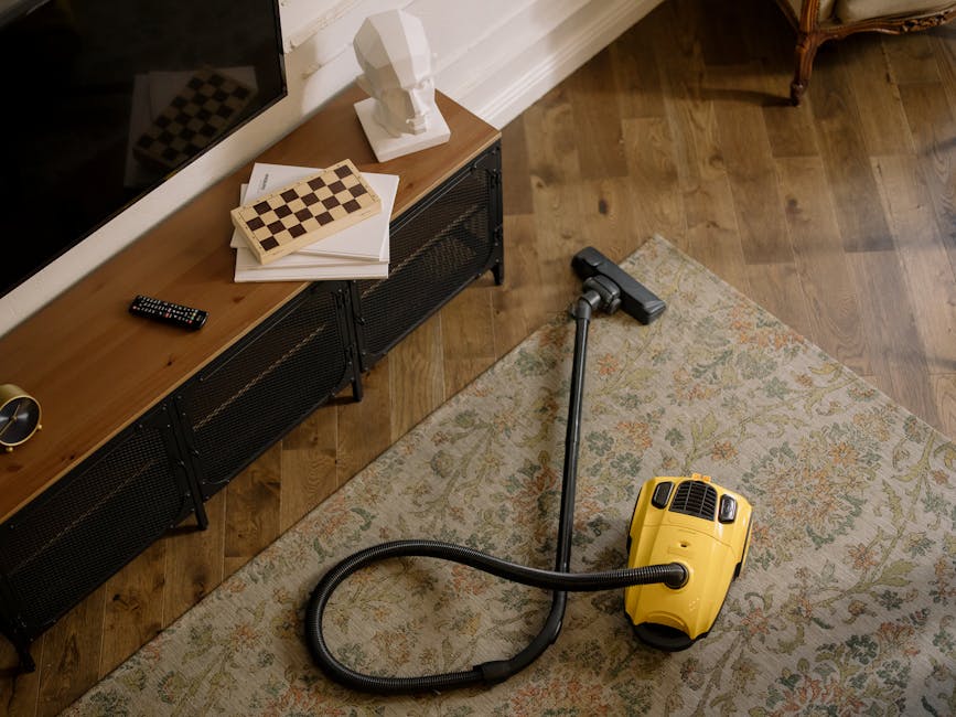 A yellow handheld vacuum cleaner with a long hose attachment is placed on a floral patterned area rug in a living room. The room features hardwood flooring, and a wooden TV console with black metal legs is positioned against the white wall, holding several books, a decorative white bust, a black remote control, and a small gold-colored object. Natural light illuminates the space, highlighting the clean and well-maintained surfaces. The scene reflects domestic surface cleaning practices, with a focus on vacuuming the rug to maintain hygiene and cleanliness, as performed by Fulham Carpet Cleaning.