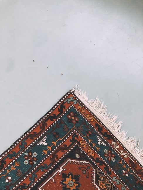 Close-up of a decorative traditional rug with geometric patterns in shades of red, blue, and beige, partially folded over a smooth, white surface with faint marks and scratches. The rug has fringed edges visible at the top right corner and is placed on a clean, well-lit area, suggesting a setting involving surface cleaning or maintenance. The image emphasizes the intricate design and textile quality, aligning with deep cleaning or surface sanitisation services offered by Fulham Carpet Cleaning for residential or commercial spaces, as referenced on the Sands End rug cleaning and stain removal SW6 page.